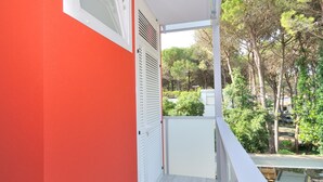 A balcony with a red wall, white shutters, and a view of trees and a parked car. - Apartment in Bibione Near the Beach (San Michele al Tagliamento)