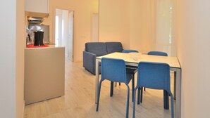 A compact kitchen with a coffee machine, a dining area with a table and chairs, and a door leading to another room. - Apartment in Bibione Near the Beach (San Michele al Tagliamento)