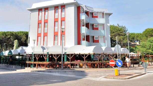 A multi-story building with a white facade and red accents, featuring balconies and a covered outdoor seating area. - Apartment in Bibione Near the Beach (San Michele al Tagliamento)