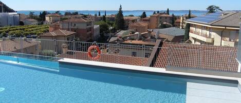 Une piscine équipée d’une bouée de sauvetage, avec une eau d’un bleu limpide, et une vue sur une ville côtière aux bâtiments aux toits de tuiles.