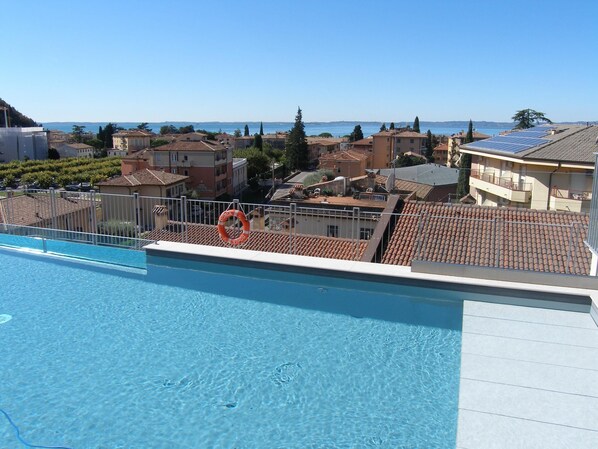 En swimmingpool med en redningskrans, krystalklart blåt vand og udsigt over en kystby med terracotta-tag.