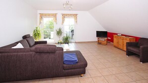 Ein Wohnzimmer mit einer Couch, einem Fernseher, einem Schrank und zwei Fenstern mit Vorhängen.