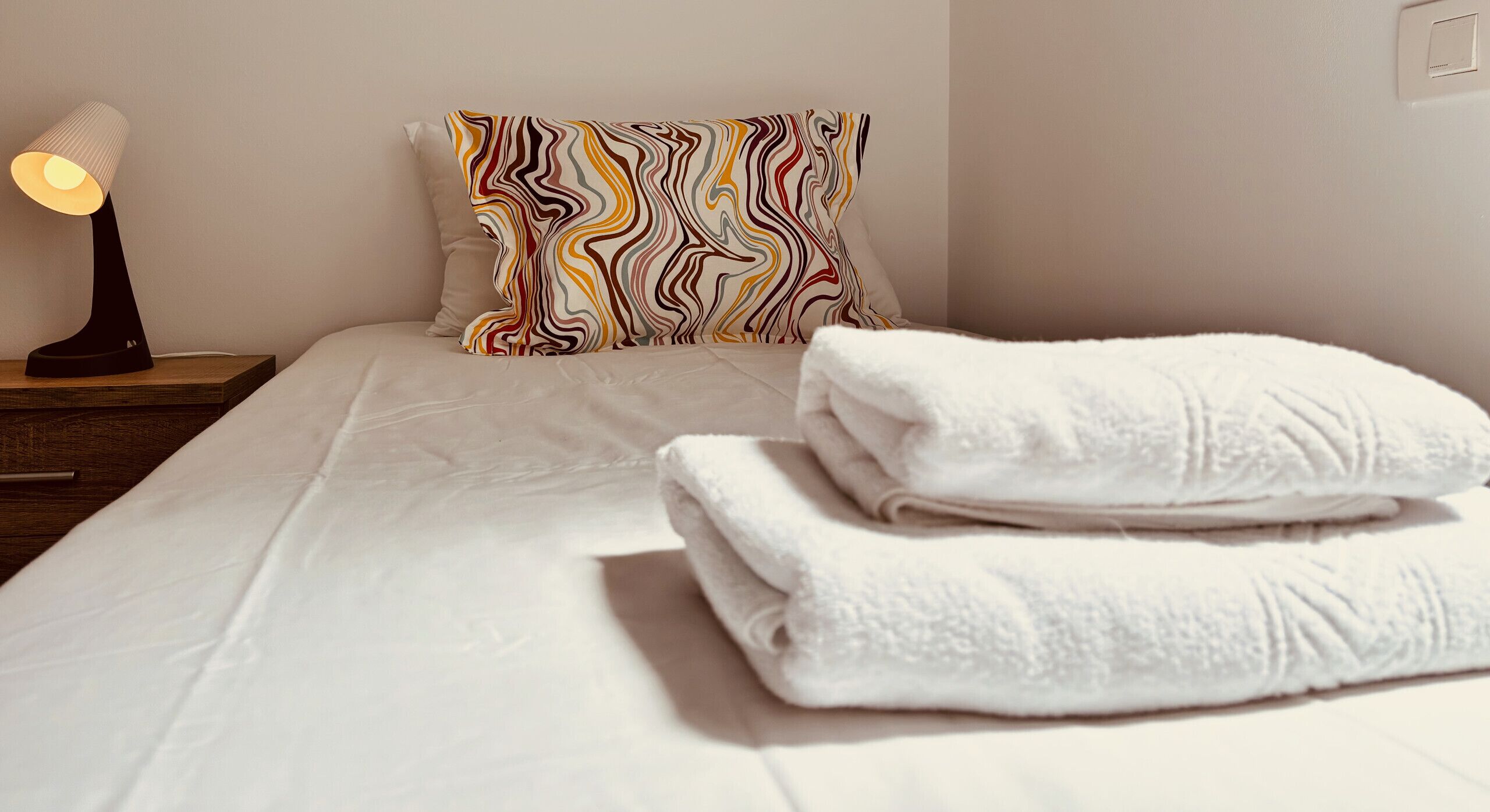 Un lit bien fait, avec des draps blancs, un coussin coloré et une lampe de chevet.