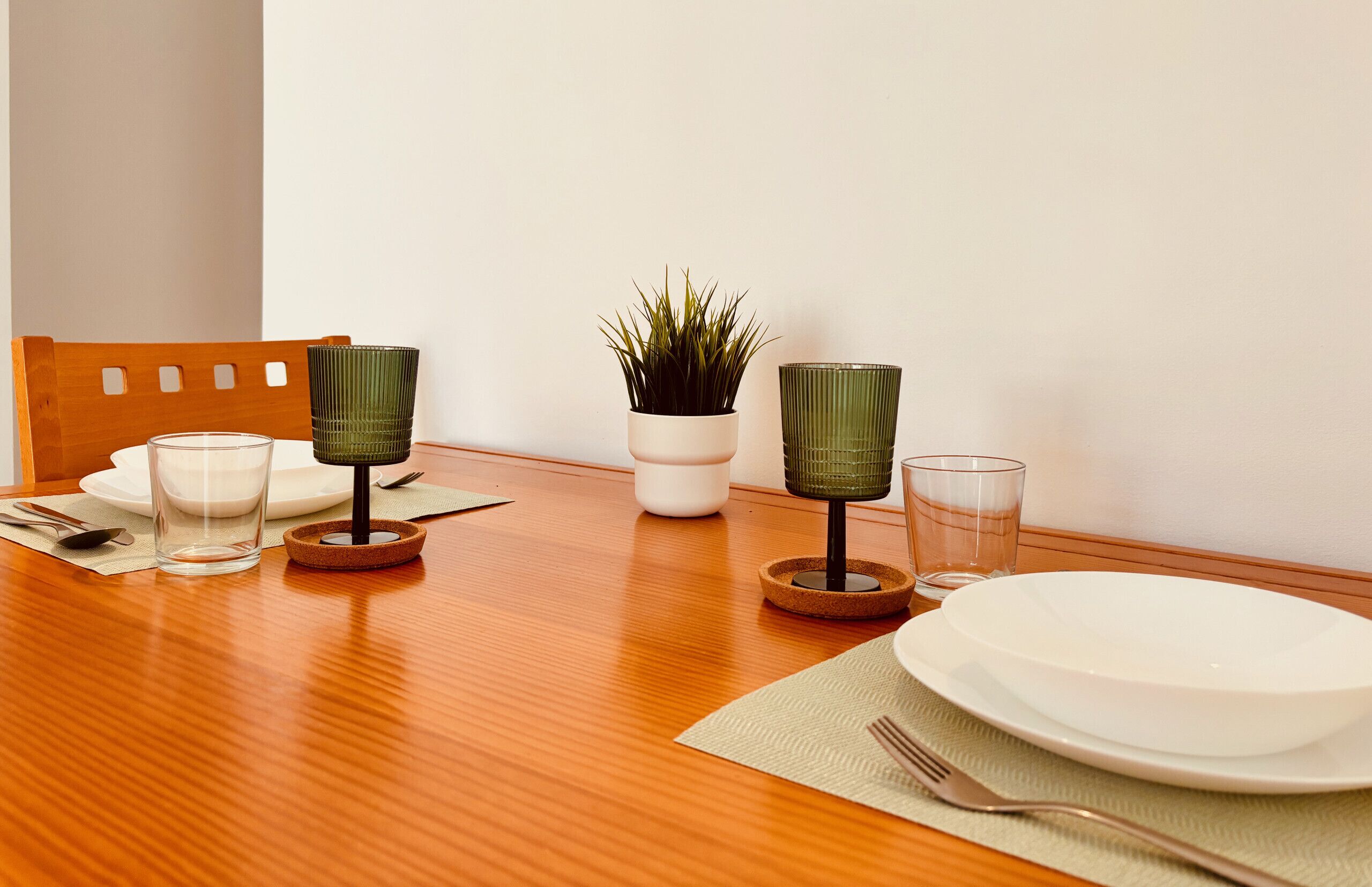 A wooden dining table with a white plate, two green glass candle holders, a potted plant, and a green glass cup.