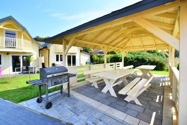 Un patio exterior con una pérgola de madera, mesas de picnic y una parrilla.
