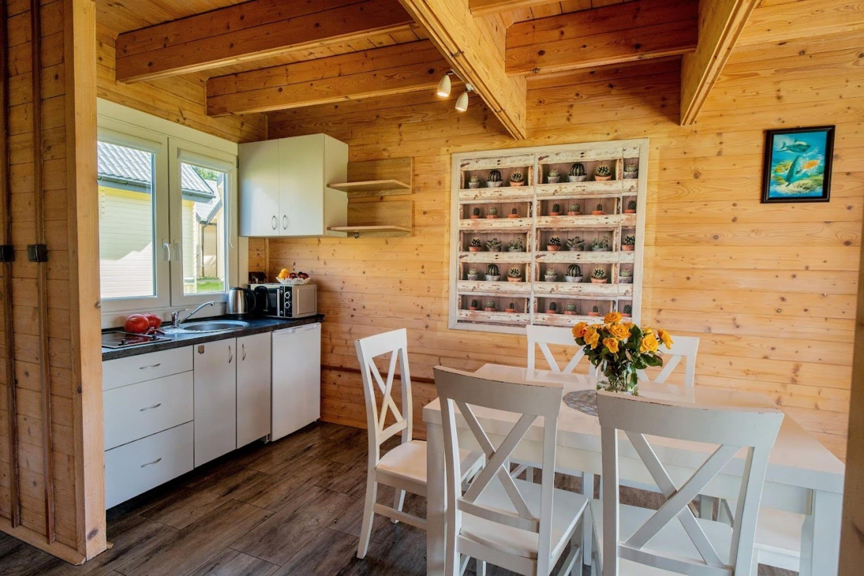 A wooden interior with a kitchen area, white cabinets, a dining table with chairs, and a wall-mounted shelf with potted plants.