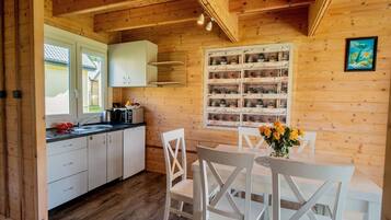 A wooden interior with a kitchen area, white cabinets, a dining table with chairs, and a wall-mounted shelf with potted plants.