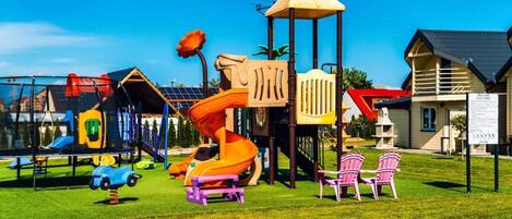 A playground with slides and climbing structures, surrounded by grass and a few chairs.