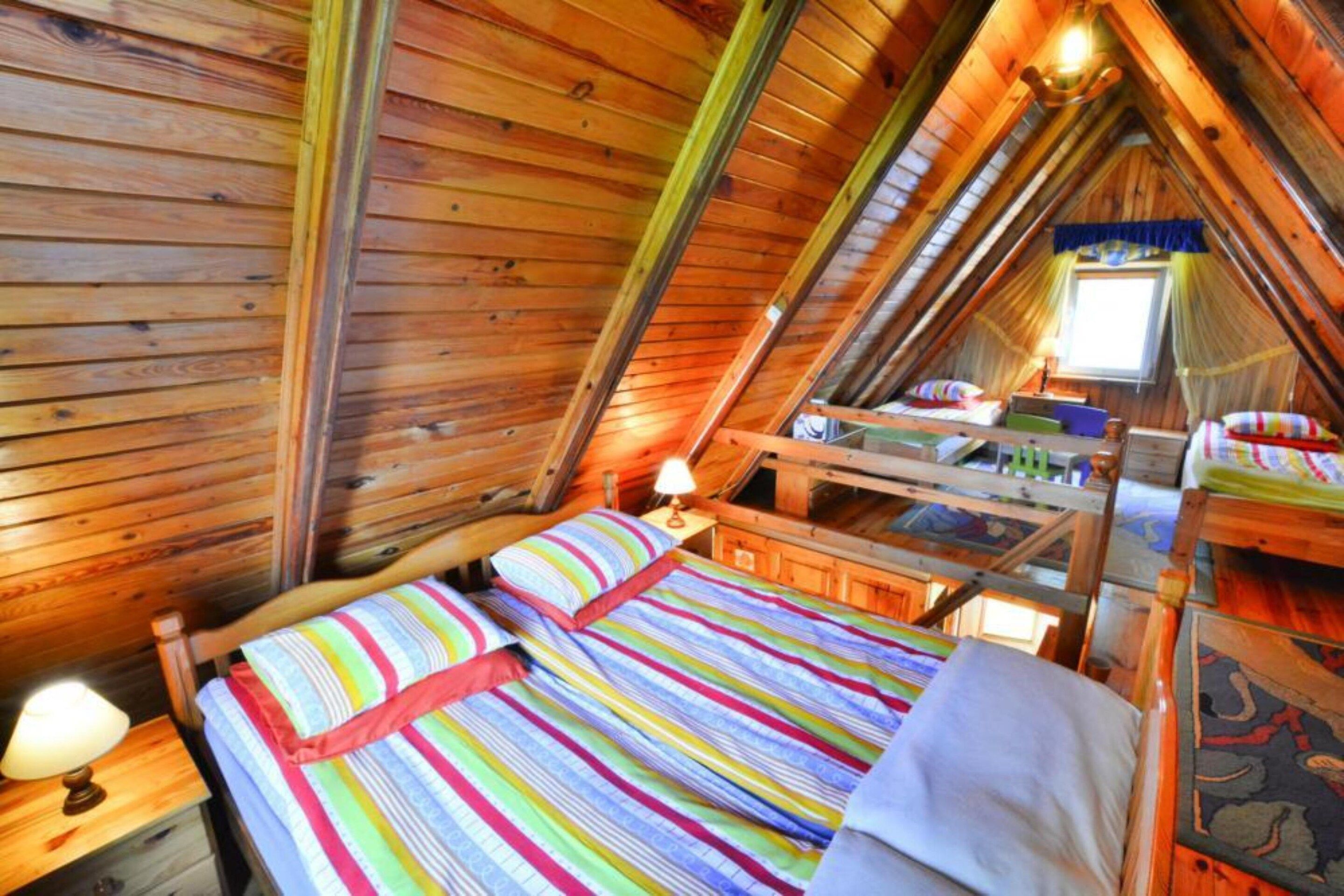 A cozy attic bedroom with a wooden sloped ceiling, a bed with colorful striped bedding, a desk with a lamp, and a window with curtains.