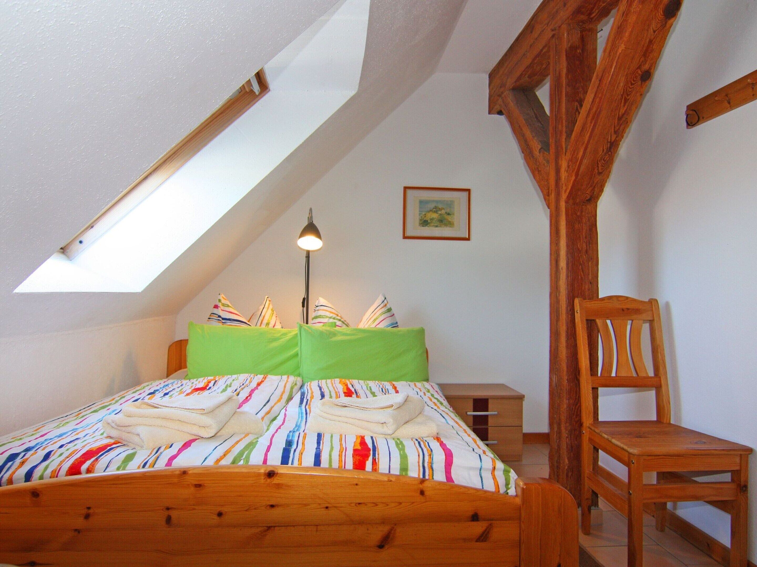 A bedroom with a sloped ceiling, a bed with colorful bedding, a bedside table, a chair, and a framed picture on the wall.