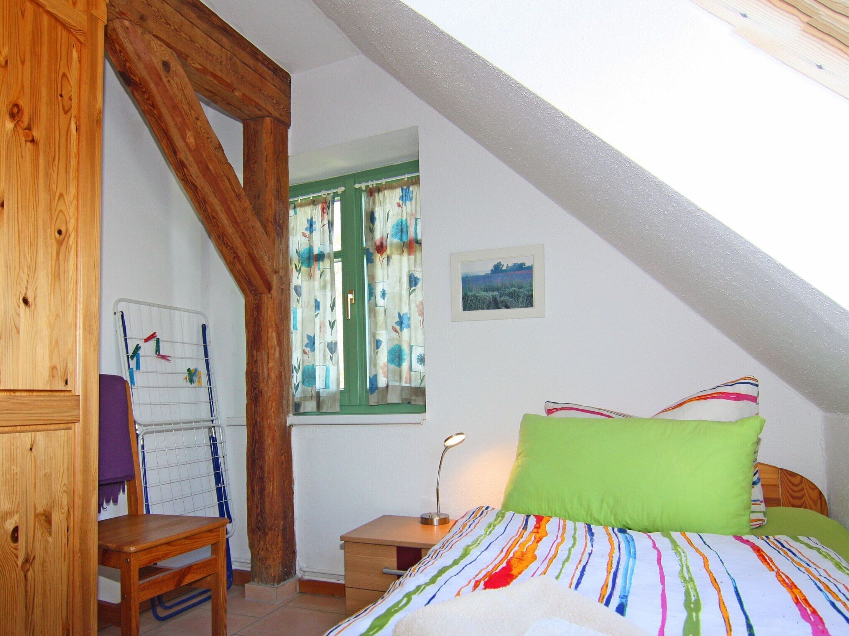 A bedroom with a sloped ceiling, a wooden chair, a desk, a bed with colorful bedding, a window with floral curtains, and a small bedside table with a lamp.