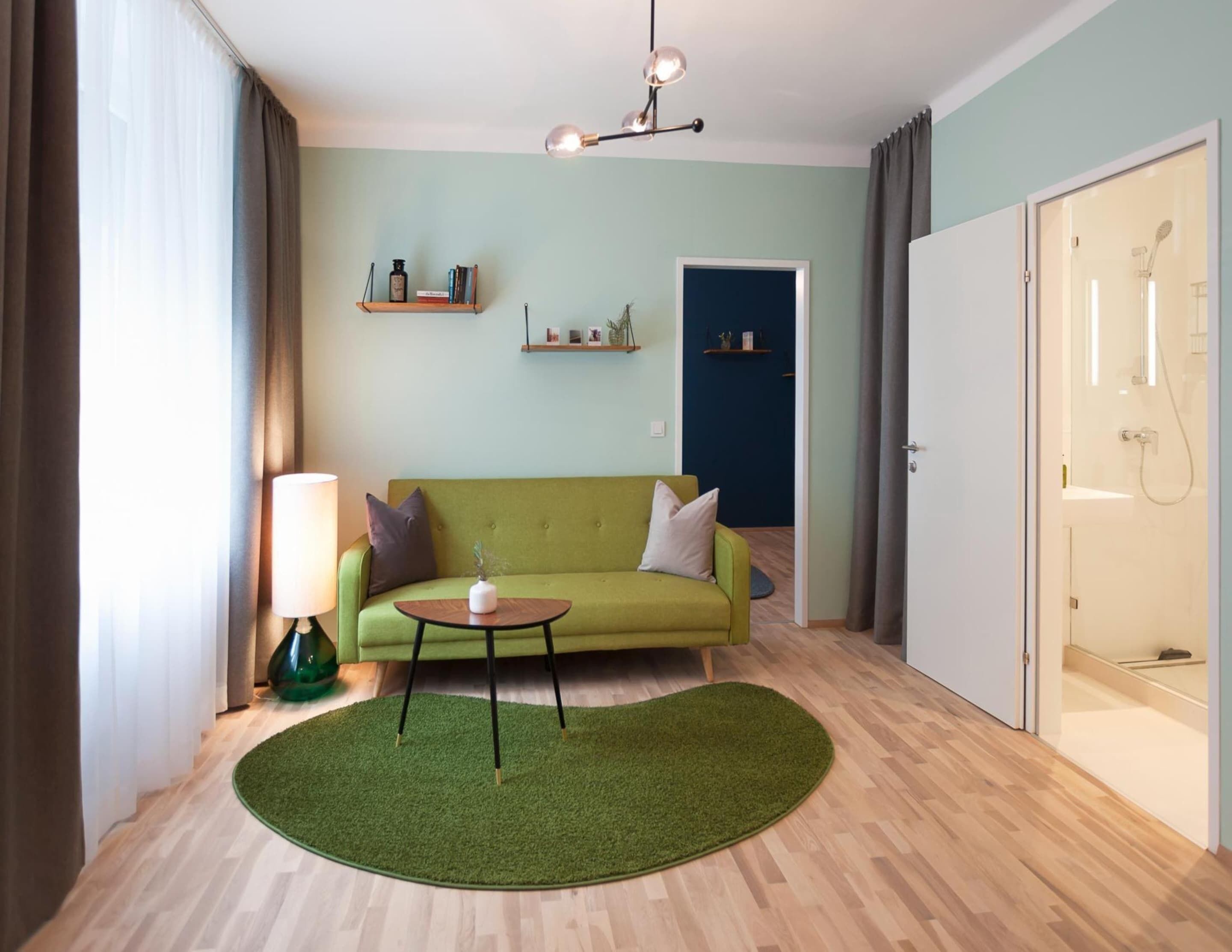 A modern living room with a green sofa, a small round table, and a green rug. There are shelves with decorative items on the wall, and a door leading to a bathroom.