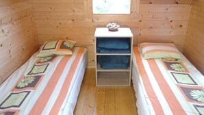 Ein kleines Zimmer mit Holzwänden und zwei Einzelbetten, jeweils mit gemusterten Bettdecken und Kissen. Eine kleine Regaleinheit mit gefalteten Handtüchern befindet sich in der Mitte des Raumes.