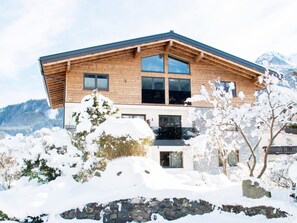A two-story wooden house with large windows, a stone foundation, and a snowy landscape with mountains in the background. - Apartment in Bramberg Near Cable Car (Bramberg am Wildkogel)