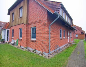 A two-story brick house with a sloped roof, a front porch, and a small front yard with a bench and a hose. - Wohnung in Wenkendorf mit Garten (Fehmarn)