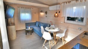 Une pièce confortable, aménagée dans le style d’un chalet, avec un plafond en bois, un canapé, une table à manger avec des chaises, un téléviseur fixé au mur et une fenêtre avec des rideaux.