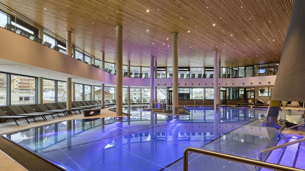 A modern indoor swimming pool with a glass barrier, multiple seating areas, and a high ceiling with a wooden design. - Apartment in Ischgl at the ski bus Stop (Ischgl)