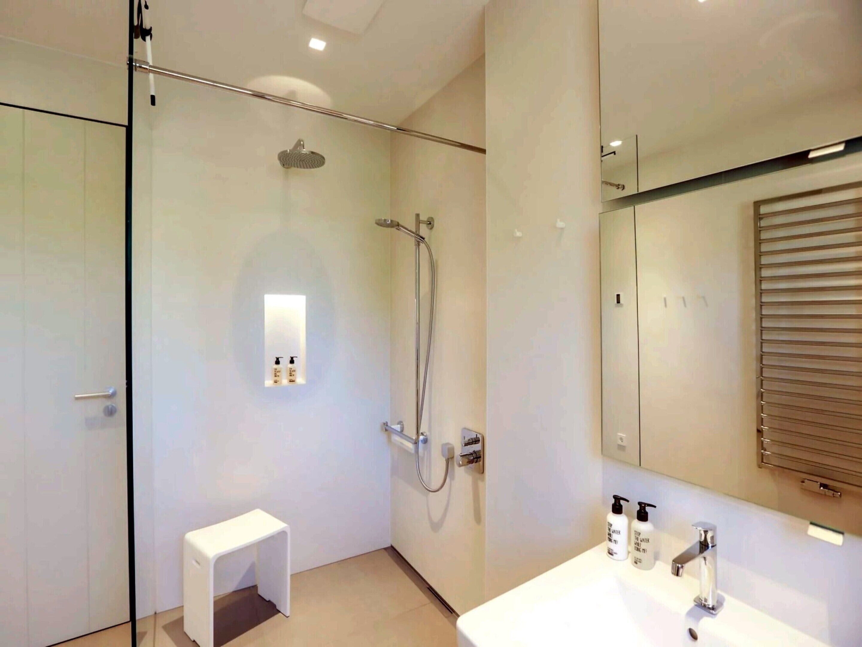 A modern bathroom with a glass shower enclosure, a small white bench, a sink with two bottles of shampoo, and a large mirror.