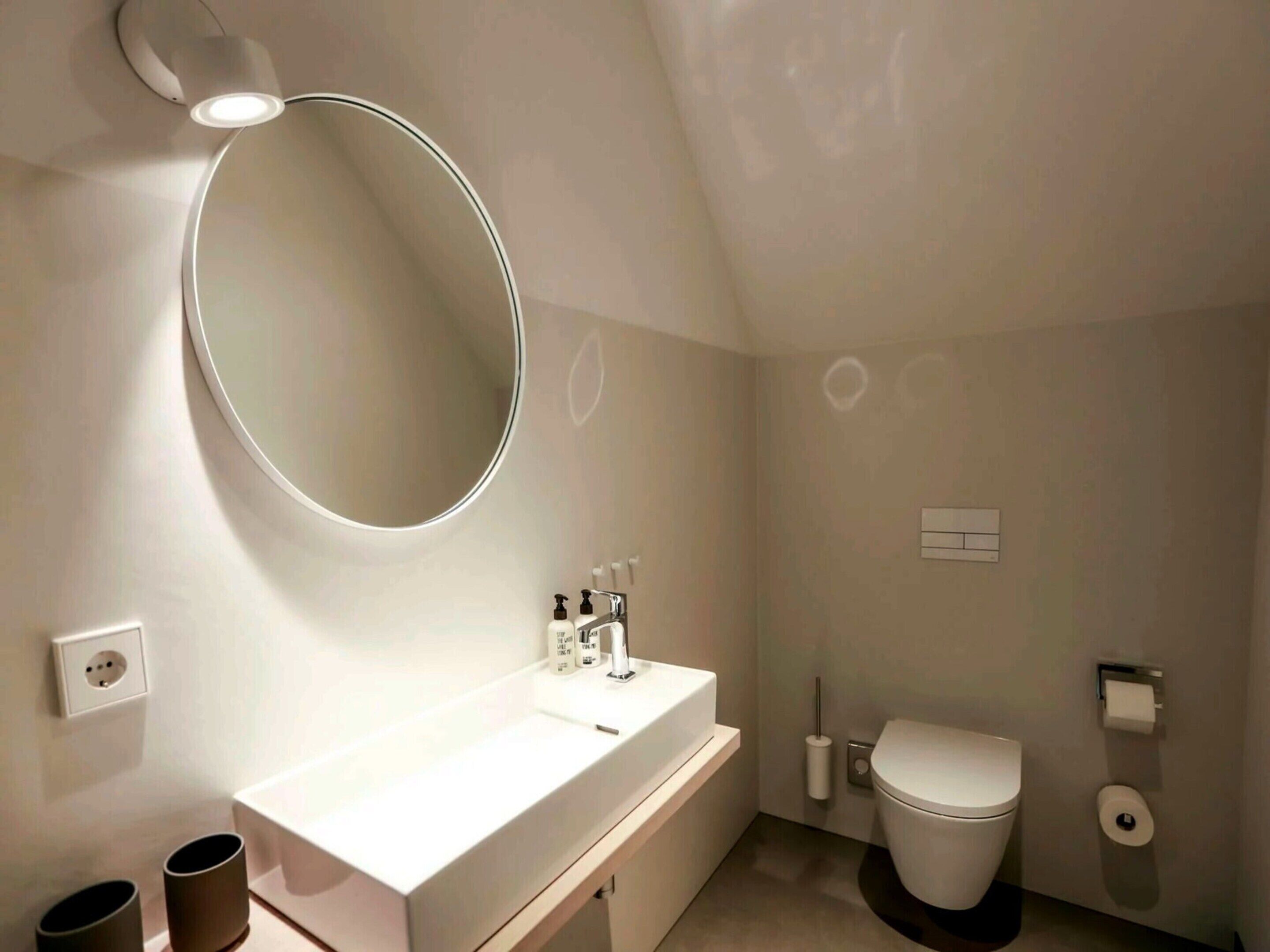 A modern bathroom with a round mirror, white sink, and toilet.