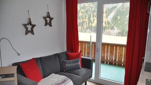 A living room with a grey sofa, red and grey cushions, a white blanket with 'Gipfelluck' text, wooden flooring, a wooden deck outside, and two star decorations on the wall. - Chalet in Steeg Near Ski Slopes (Steeg)