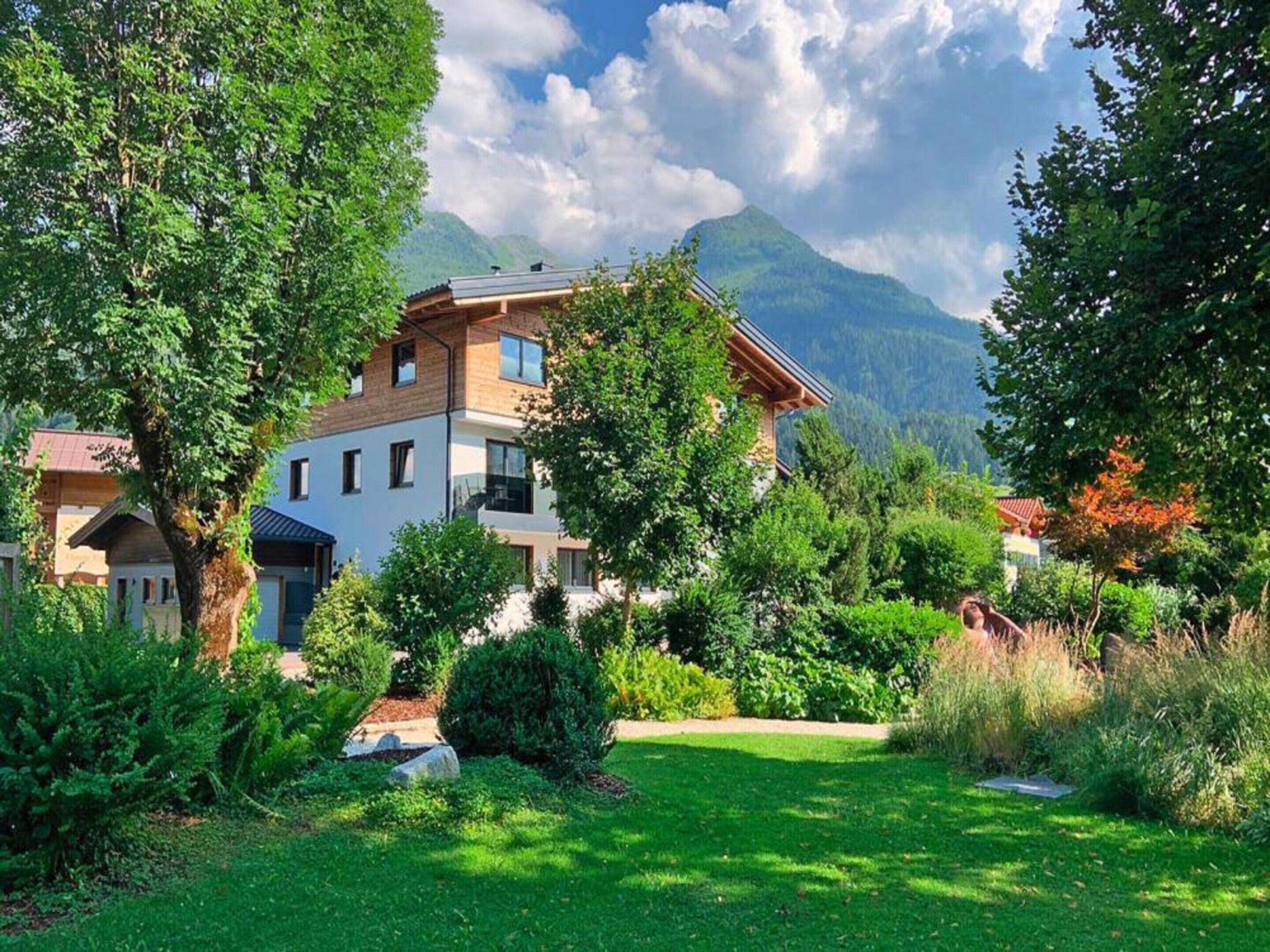 A two-story house with a garden in front, surrounded by trees and mountains in the background.