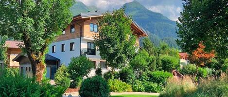 A two-story house with a garden in front, surrounded by trees and mountains in the background.