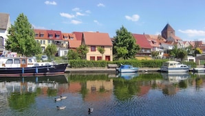 Un río tranquilo con barcos amarrados a la izquierda, rodeado de casas tradicionales con tejados rojos y una torre de iglesia al fondo.