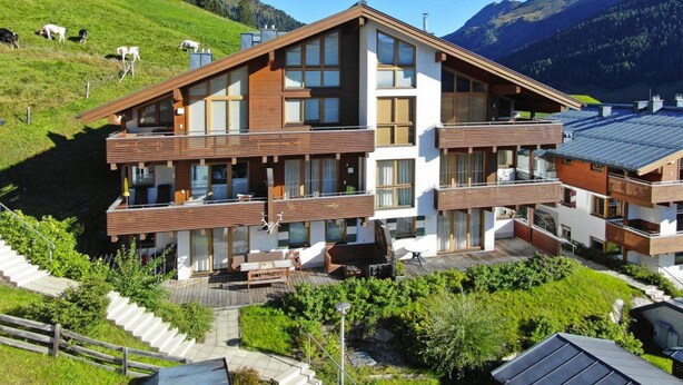 Un bâtiment moderne à deux étages, doté de grandes fenêtres et d’une façade en bois, entouré d’une végétation luxuriante et adossé à un paysage montagneux.