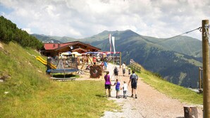 Un chalet de montagne doté d’un terrain de jeux, d’un sentier de terre menant à celui-ci et offrant une vue sur les montagnes environnantes.