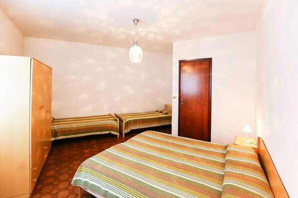 A small, single-room hotel bedroom with a wooden wardrobe, a bed with a striped bedspread, two bedside tables, a nightstand with a lamp, and a wooden door. - Holiday Village Tivoli in Bibione (San Michele al Tagliamento)