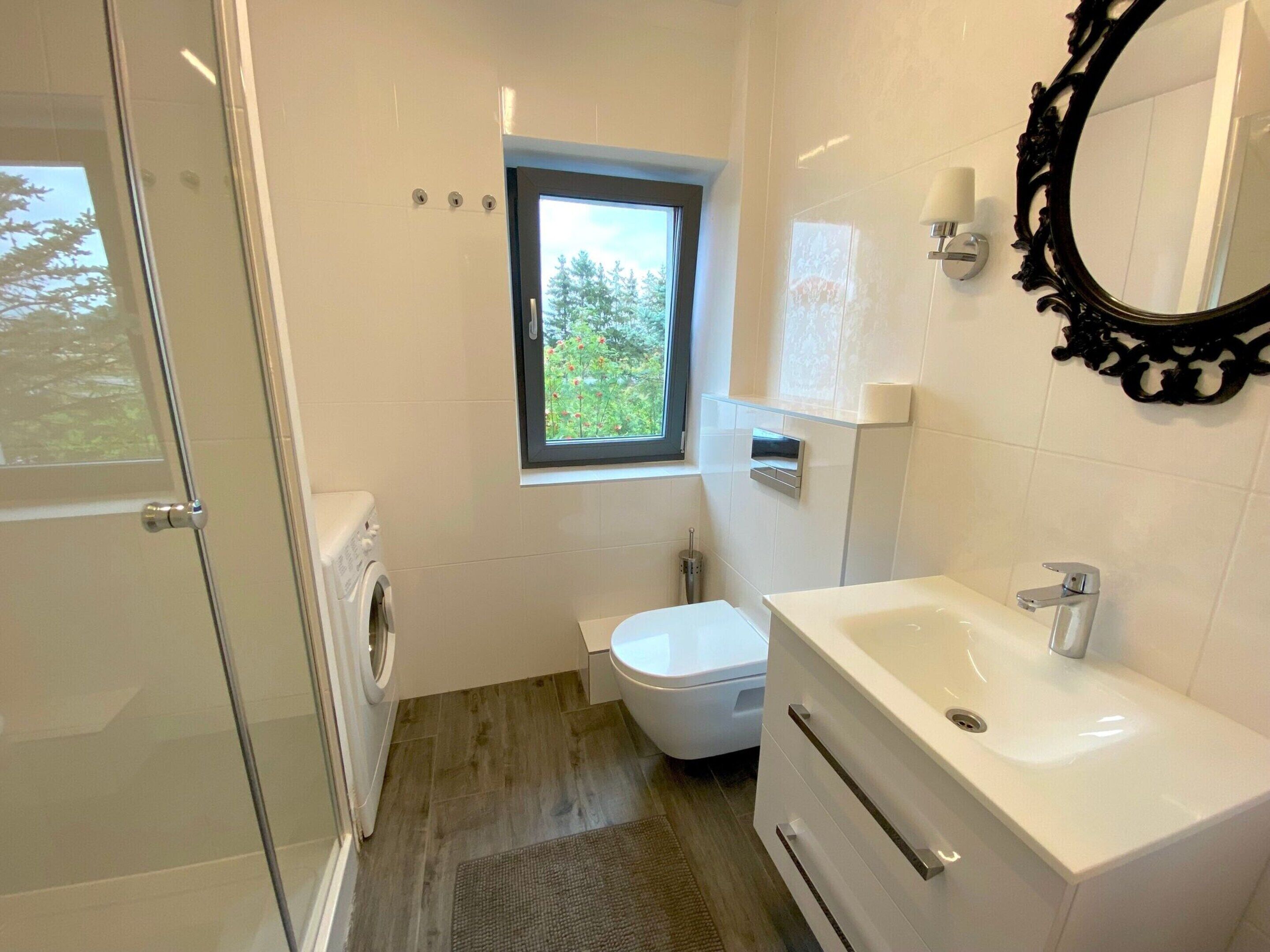 A modern bathroom with a glass shower door, a toilet, a sink, and a window with a view of greenery.