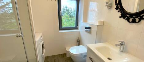 A modern bathroom with a glass shower door, a toilet, a sink, and a window with a view of greenery.