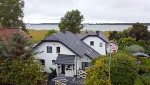 Une maison à deux étages, avec un toit en pente, un porche à l’avant et une vue sur un lac.