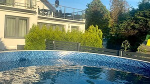 Une piscine avec un bord carrelé, un balcon agrémenté de mobilier d’extérieur et un bâtiment doté de grandes fenêtres et d’une antenne parabolique.