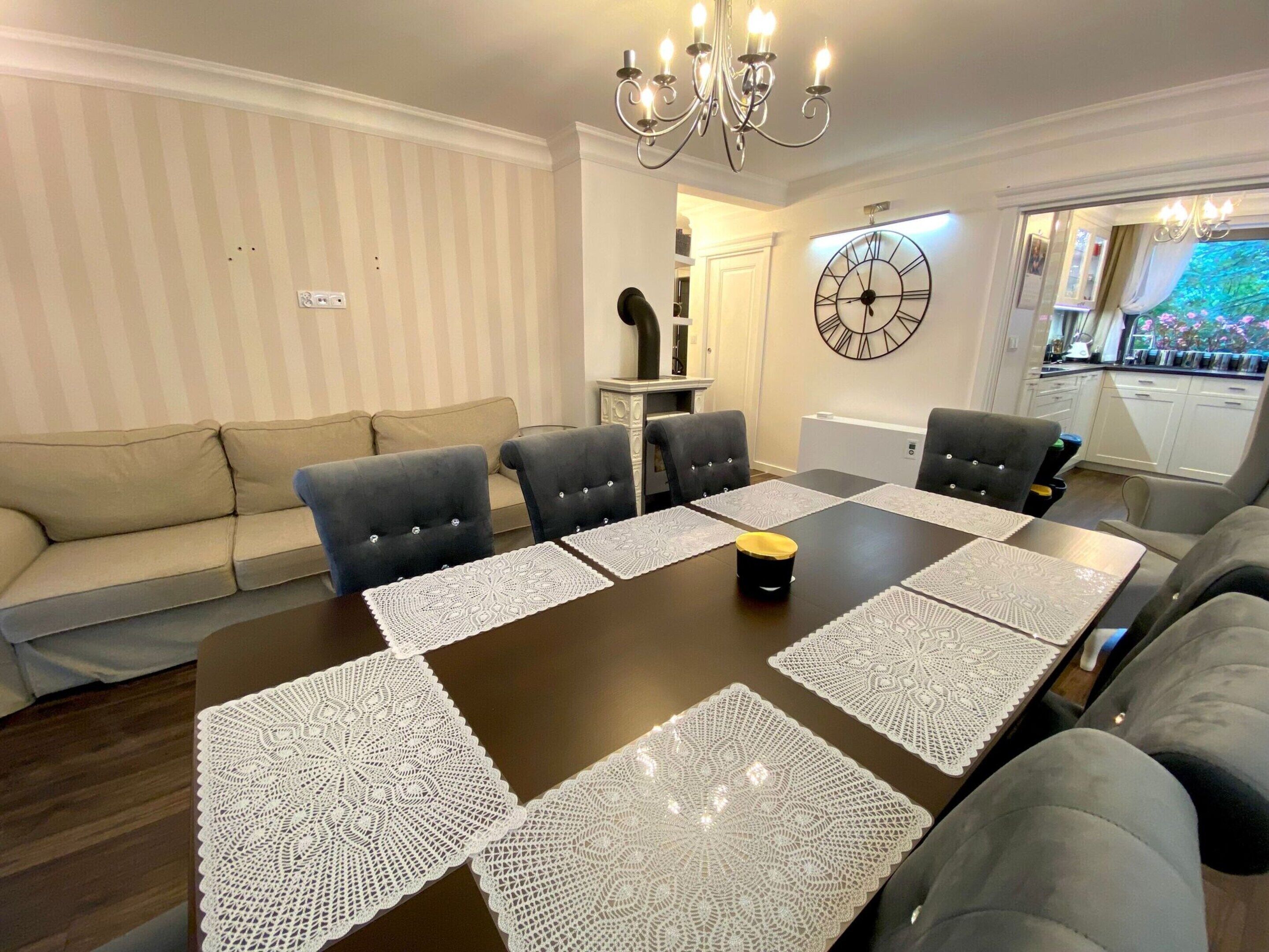 Une salle à manger avec une table en bois foncé, des sets de table en dentelle blanche et des chaises recouvertes d’un tissu gris.