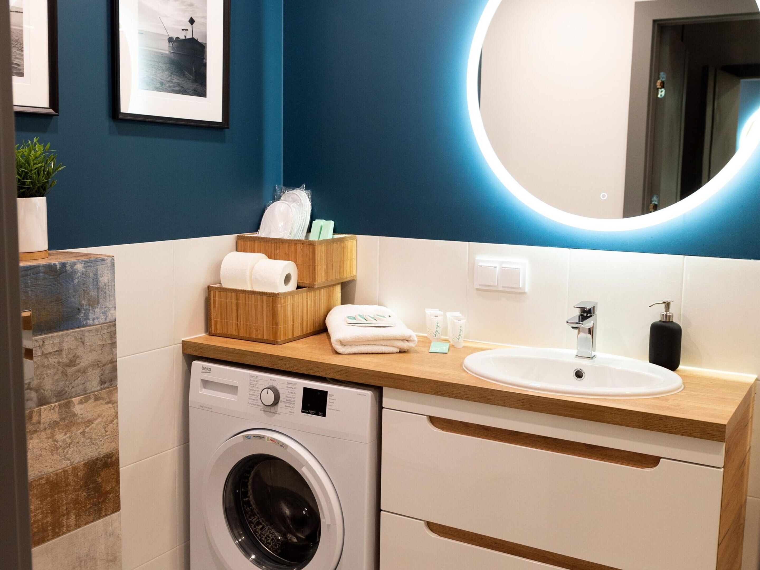 A modern bathroom with a washing machine, a sink, and a mirror. There are towels and toiletries on the counter.