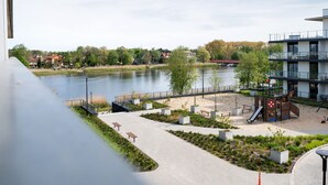 Un parco lungo il fiume con un'area giochi, panchine e un edificio moderno con facciata in vetro.