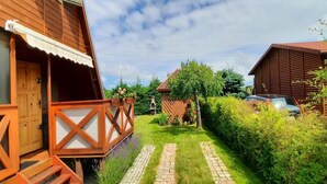 A wooden house with a porch, a paved pathway, and a car parked in the background. - Comfortable House With Private Garden, Rusinowo (Postomino)