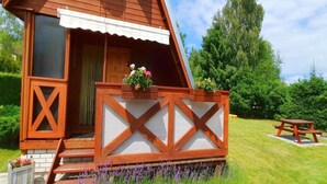 A small wooden cabin with a porch, a wooden door, and a window. There is a picnic table and a flower bed with purple flowers in front of the cabin. - Comfortable House With Private Garden, Rusinowo (Postomino)