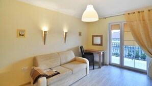 Un salon avec un canapé, une table et une chaise. Il y a des lampes fixées au mur et une fenêtre donnant sur un balcon.