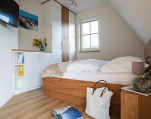 A bedroom with a wooden bed, white bedding, a bedside table with a lamp, a white wall-mounted TV, a window, and a bookshelf with books and a vase of flowers. - Luxushaus Bodden mit Sauna (Fuhlendorf)