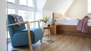 Una camera da letto con un letto in legno, una poltrona blu, un tavolino con un vaso di fiori e una finestra vista sul verde.