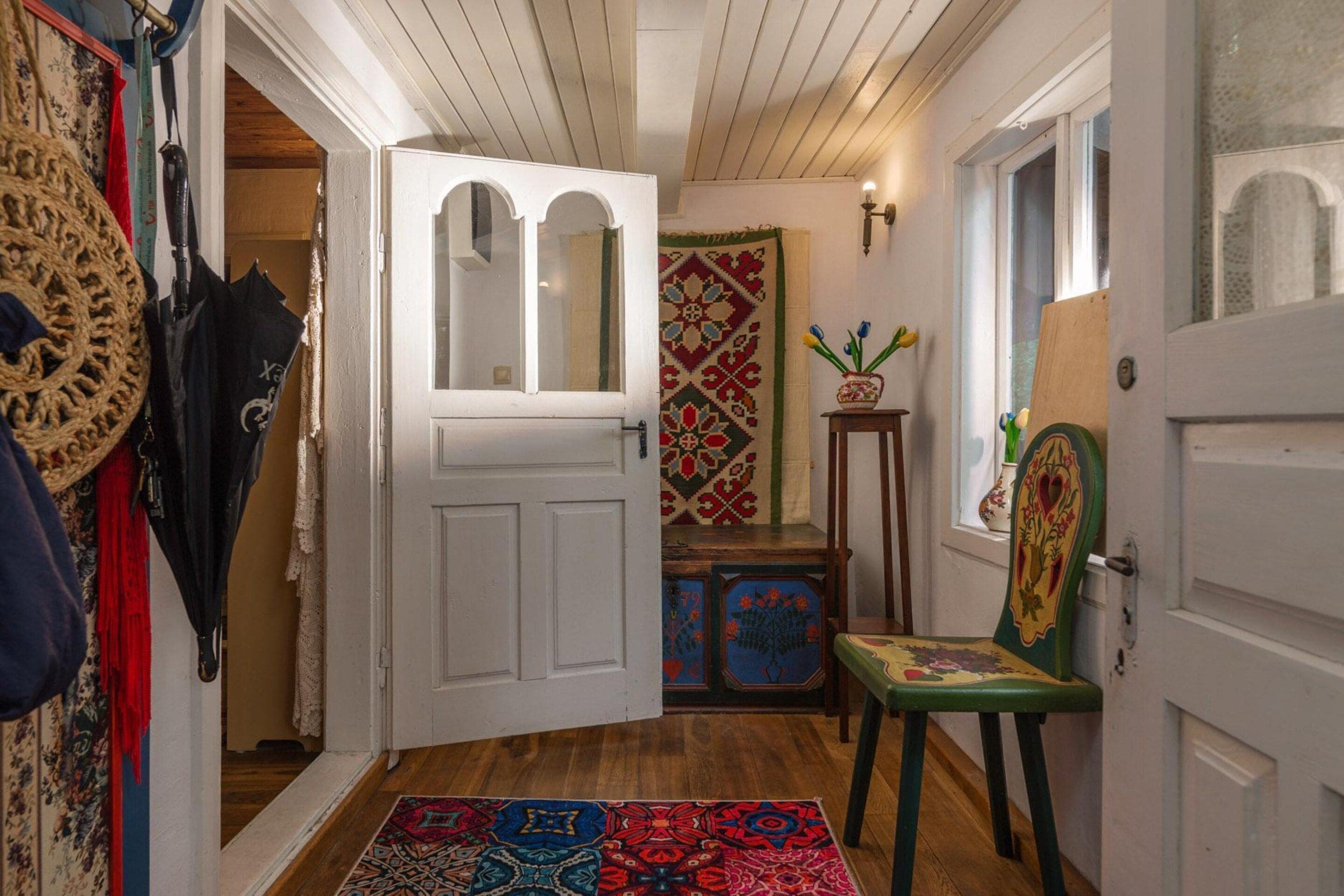 Un pasillo con una puerta blanca, un tapete estampado, una silla y una pequeña mesa con un florero.