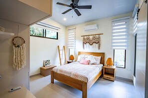 Un dormitorio con una cama de madera, un ventilador de techo, una mesita de noche con lámpara, un aire acondicionado instalado en la pared y una ventana con persianas.