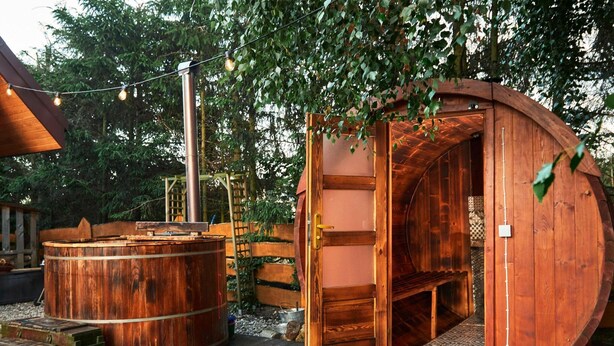 Un jacuzzi de madera y una sauna con puerta de madera, rodeados de árboles y una terraza de madera.