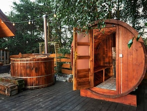 A wooden hot tub and a sauna with a wooden door, surrounded by trees and a wooden deck. - Holiday Home by Lake Zarnowieckie With Sauna (Gniewino)