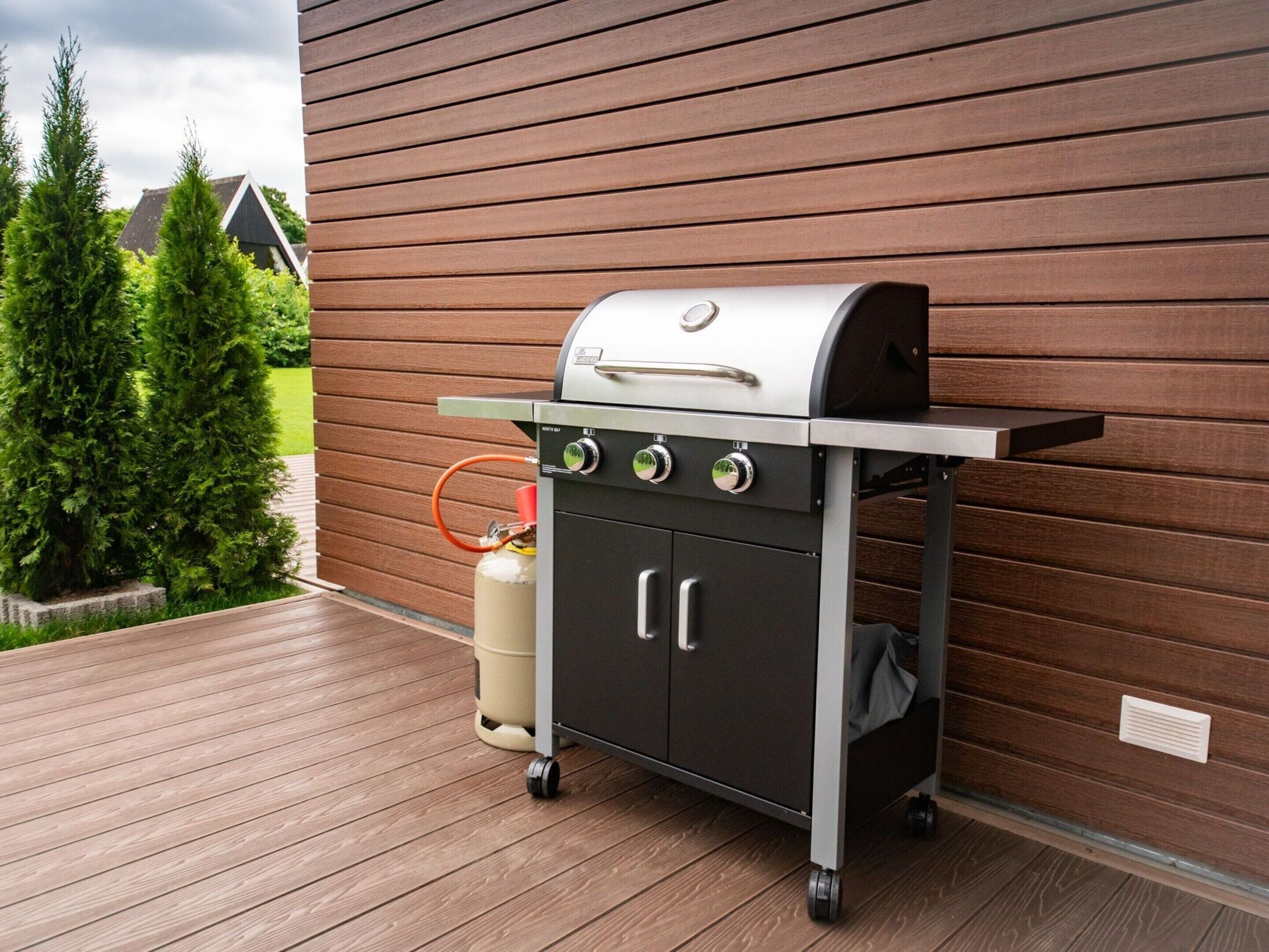 Ein tragbarer Grill mit vier Kochfeldern, einem Seitenbrenner und einem Stauraum, auf einer Holzterrasse vor einer Holzwand platziert.