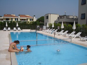 Ein Schwimmbecken mit Liegestühlen und Sonnenschirmen, umgeben von Gebäuden.