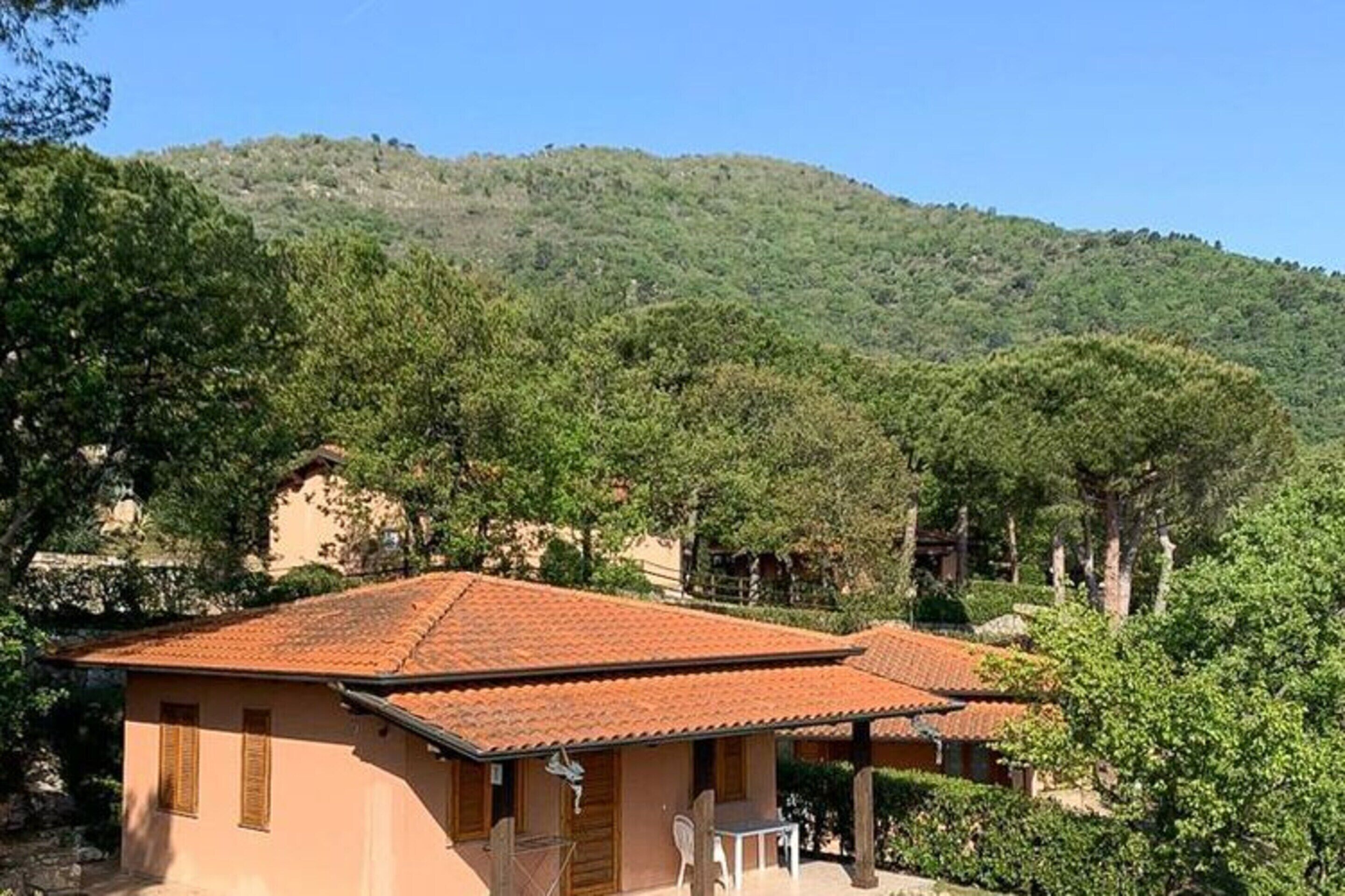 Une propriété située sur une colline, avec des bâtiments aux toits de tuiles rouges, entourée d’une végétation luxuriante et dominée par une chaîne de montagnes en arrière-plan.
