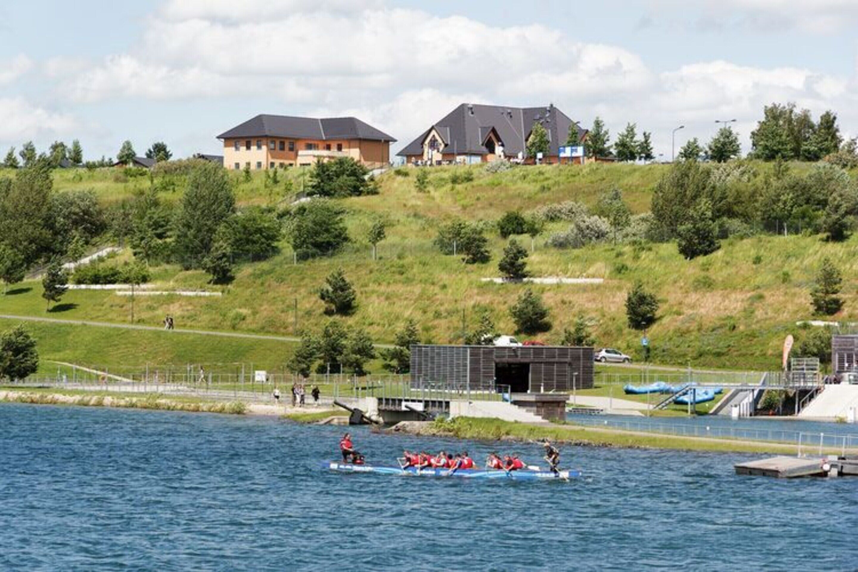 A lakeside view with a rowing team in a boat, a dock, and buildings on a hill in the background.